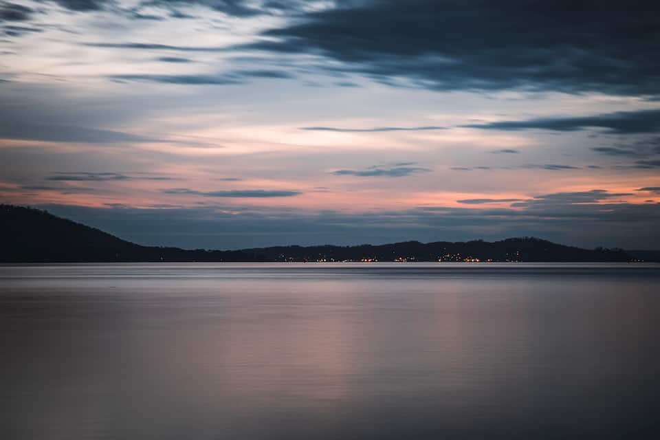 A calm lake at dusk with a mountain in the background and city lights on the horizon.