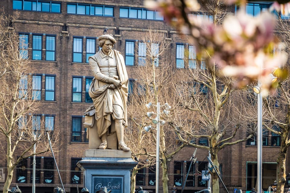 Une statue d’une personnalité historique se dresse sur une place de la ville, avec un bâtiment en briques en arrière-plan.