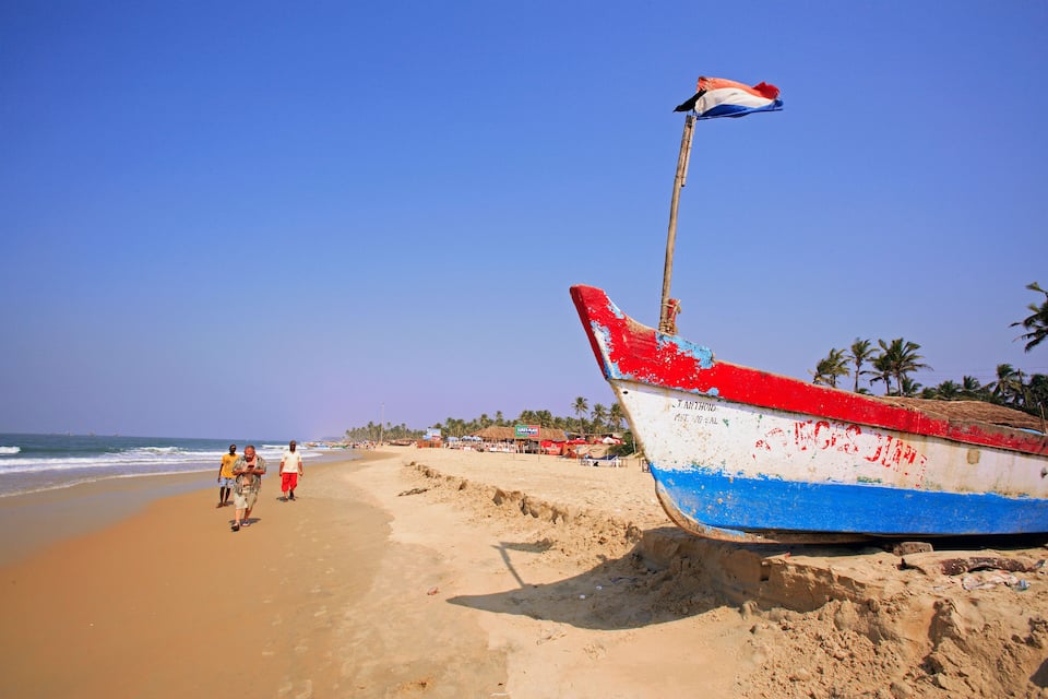 A beach with a partially submerged boat, people walking, and palm trees in the background.