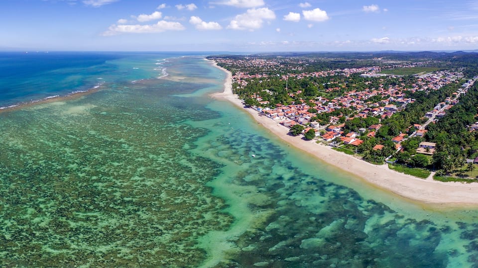 An aerial view of a coastal town with a beach, clear waters, and dense vegetation.