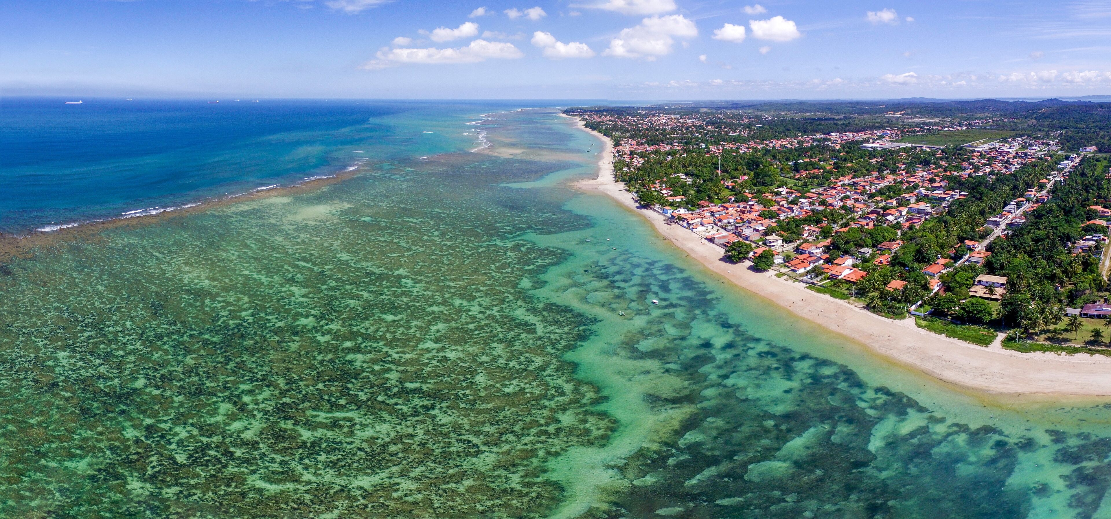 An aerial view of a coastal town with a beach, clear waters, and dense vegetation.