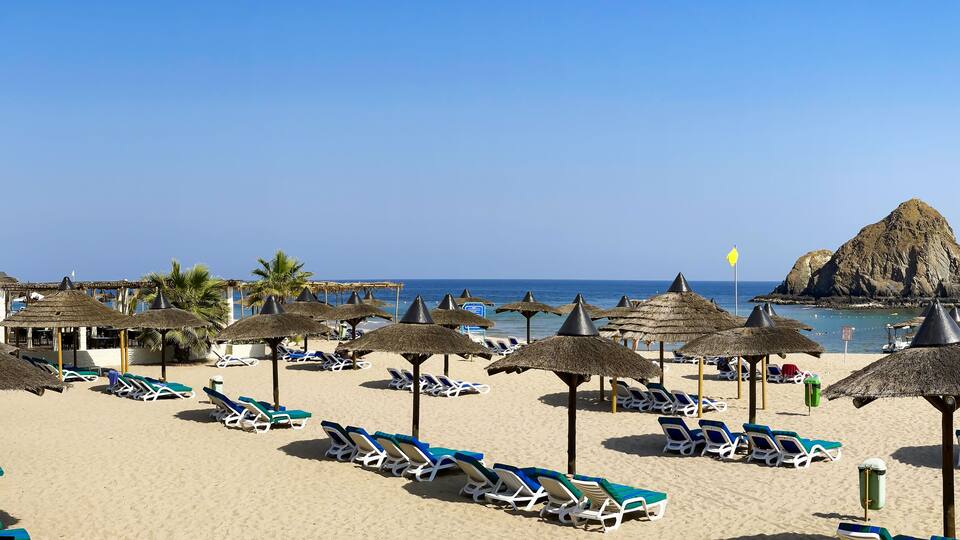 A beach with sun loungers and thatched umbrellas, palm trees, and a rocky formation in the water.
