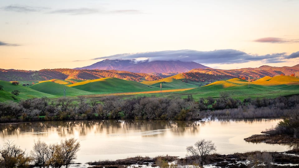 A lake reflecting a mountain range, surrounded by green fields and trees.
