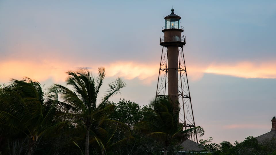 A lighthouse with a glass lantern room, surrounded by palm trees and a sunset sky.