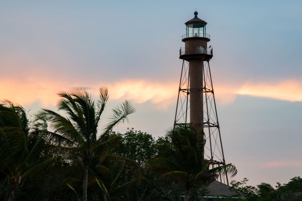 Un faro con una linterna de cristal, rodeado de palmeras y un cielo al atardecer.