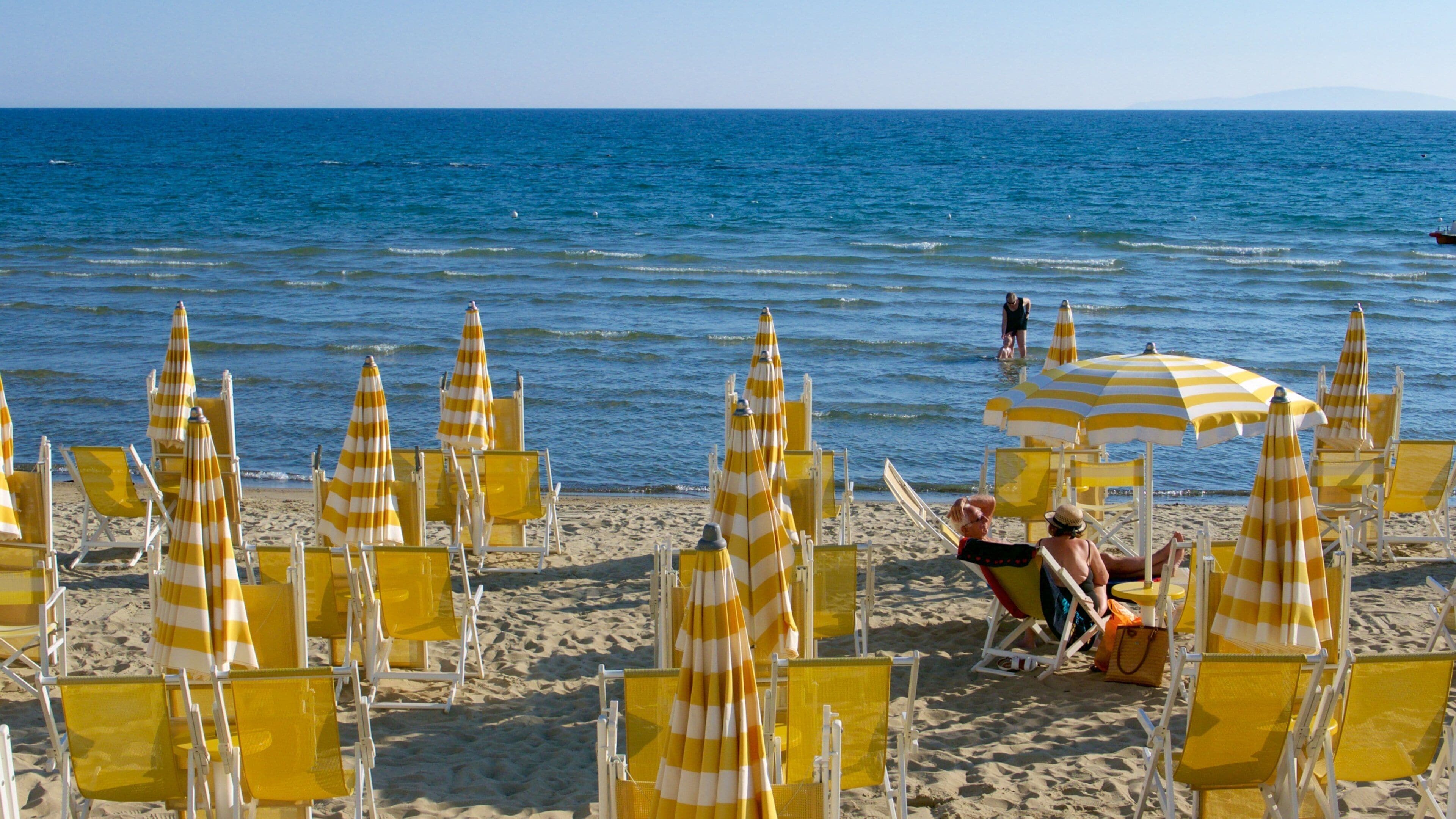 A beach with yellow striped umbrellas and chairs, a clear blue sea, and a person sunbathing.