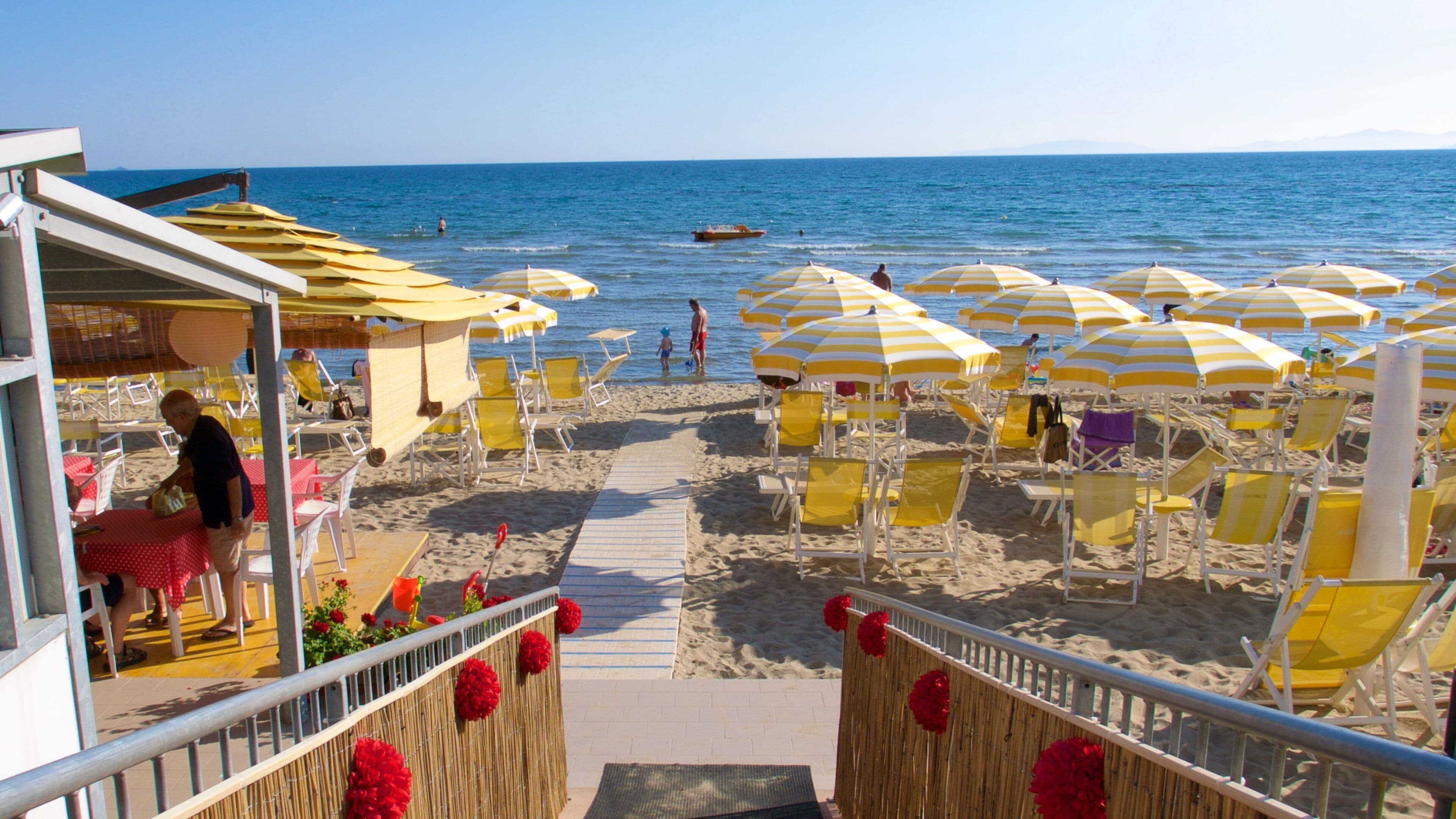 A beach with yellow and white striped umbrellas, sun loungers, and a wooden fence with red flowers.