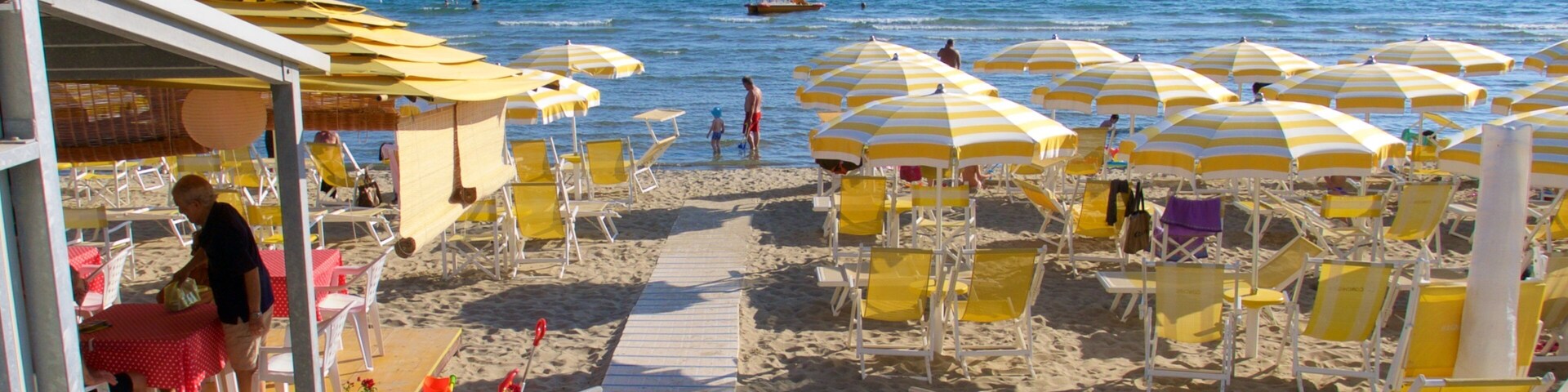 Una playa con sombrillas amarillas y blancas a rayas, tumbonas y una valla de madera adornada con flores rojas.