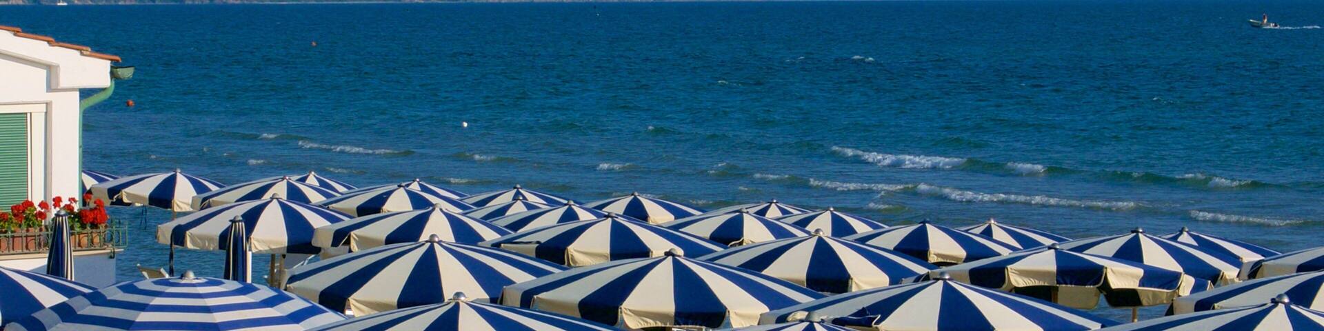 A beach with rows of striped beach umbrellas and a mountainous coastline in the background.