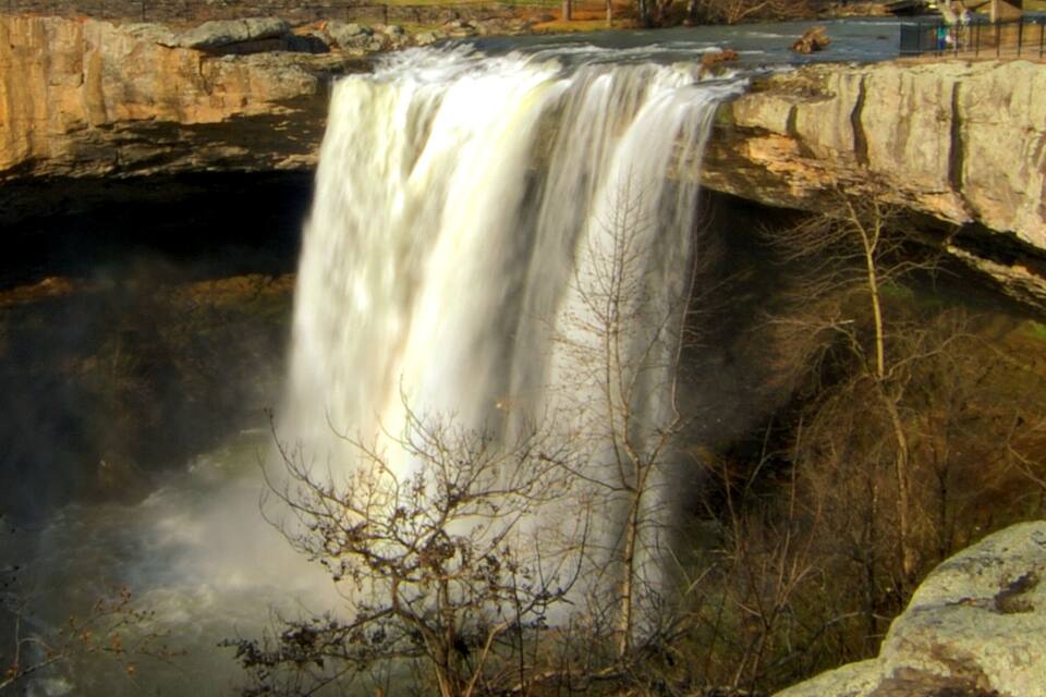 Uma cascata despenca sobre um afloramento rochoso, com uma ponte acima.