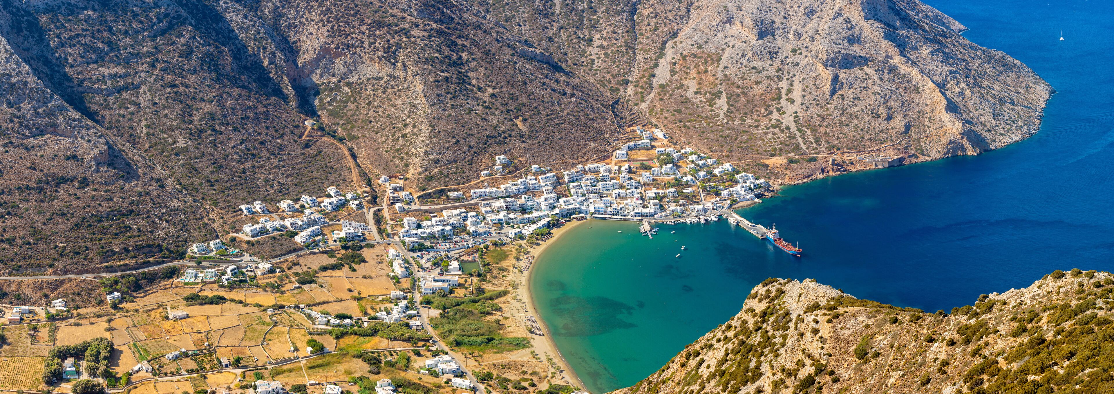 A coastal town nestled between mountains, with a clear blue sea and a winding road leading to the beach.