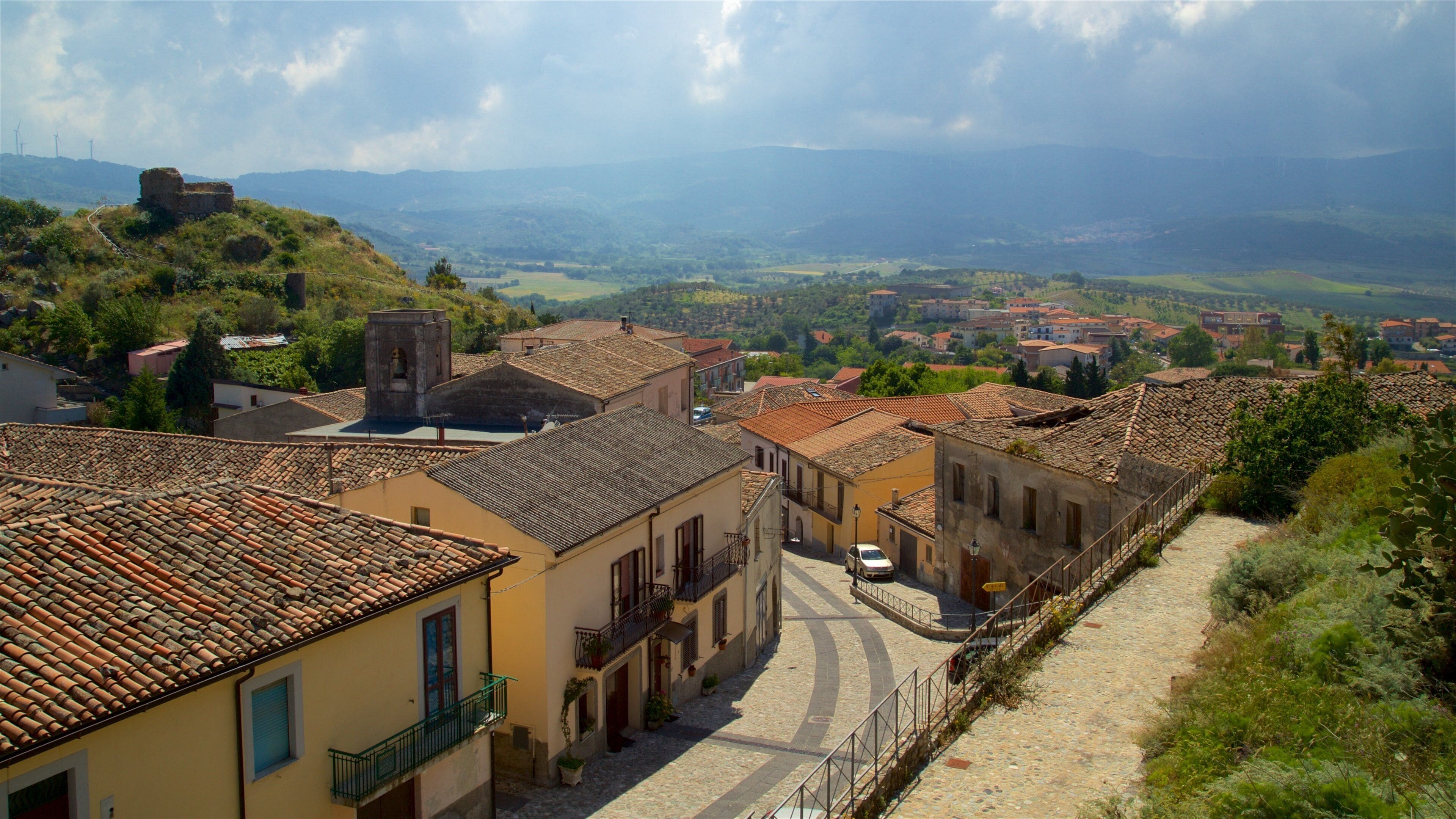 A cobblestone street in a historic town with terracotta-roofed buildings, a church, and rolling hills in the background.