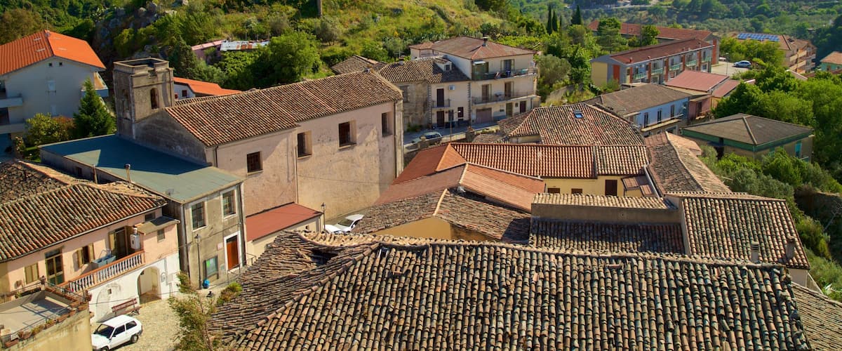 A village with terracotta-roofed buildings, a historic stone tower, and lush green hills in the background.