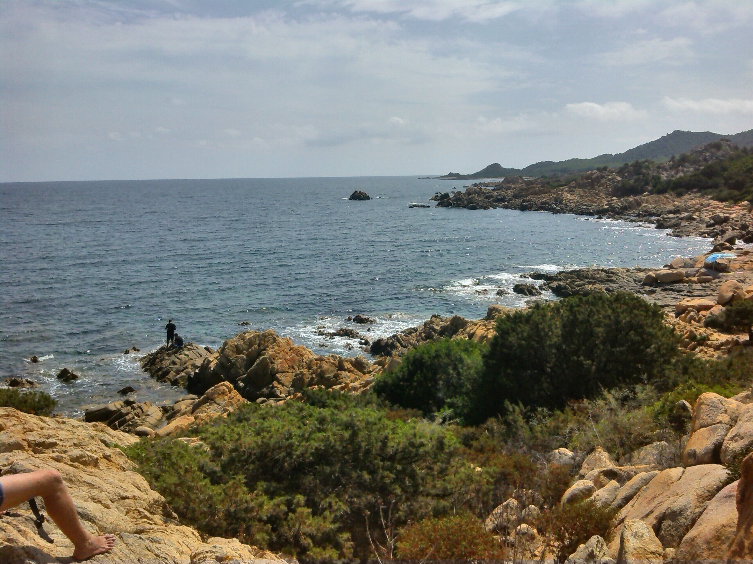 A rocky coastline with a clear view of the sea, a person standing on a rock, and a distant island.