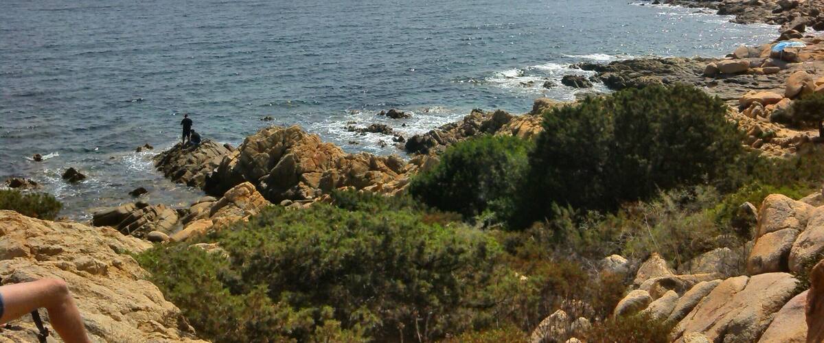 A rocky coastline with a clear view of the sea, a person standing on a rock, and a distant island.