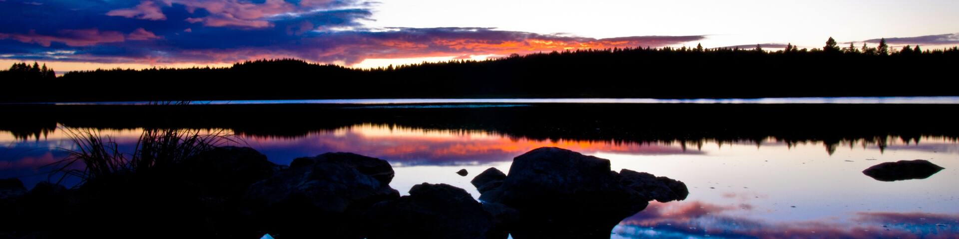 A sunset over a calm lake with silhouetted trees and a rocky shoreline.