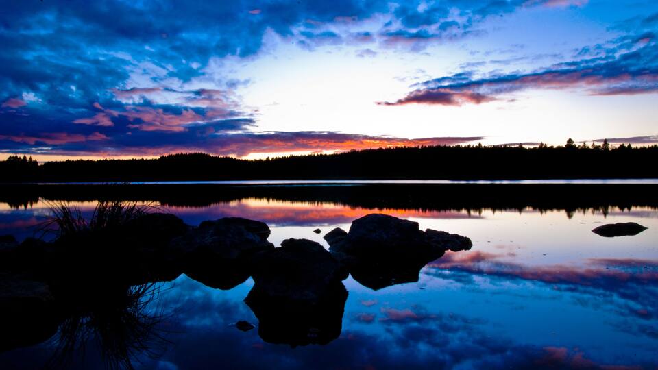 A sunset over a calm lake with silhouetted trees and a rocky shoreline.