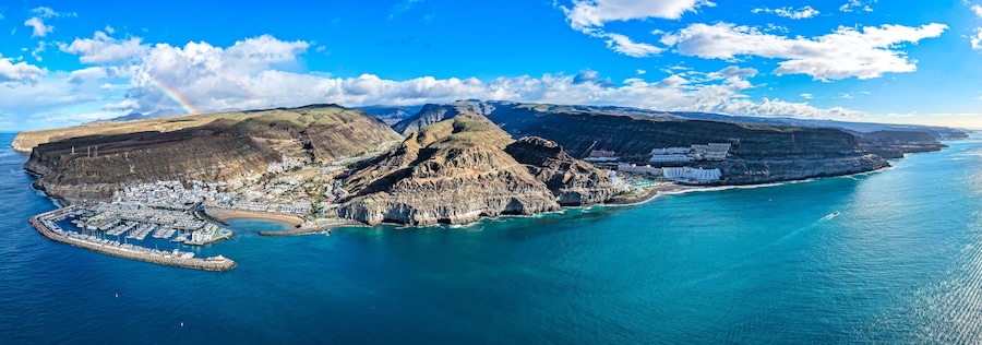 Un pueblo costero con una montaña imponente, un puerto deportivo y un cielo azul despejado.