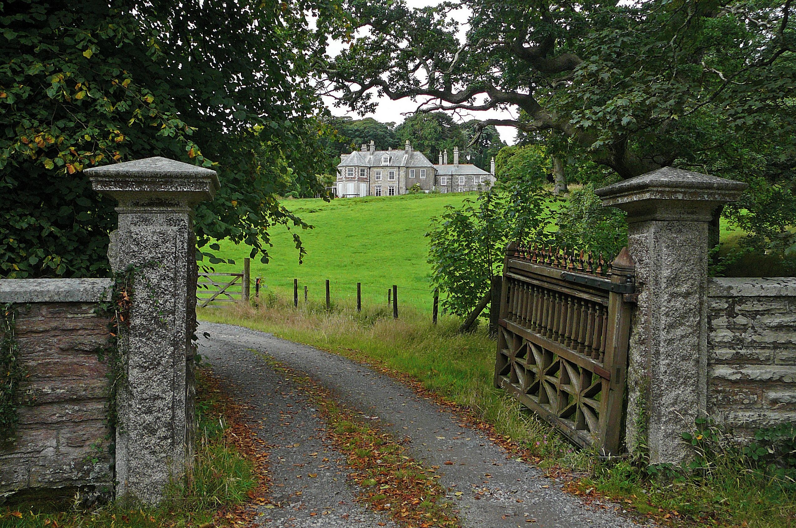 A stone gate leading to a large house on a grassy hill.