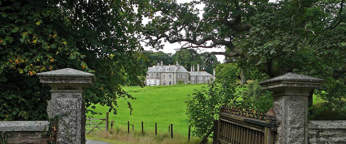 A stone gate leading to a large house on a grassy hill.