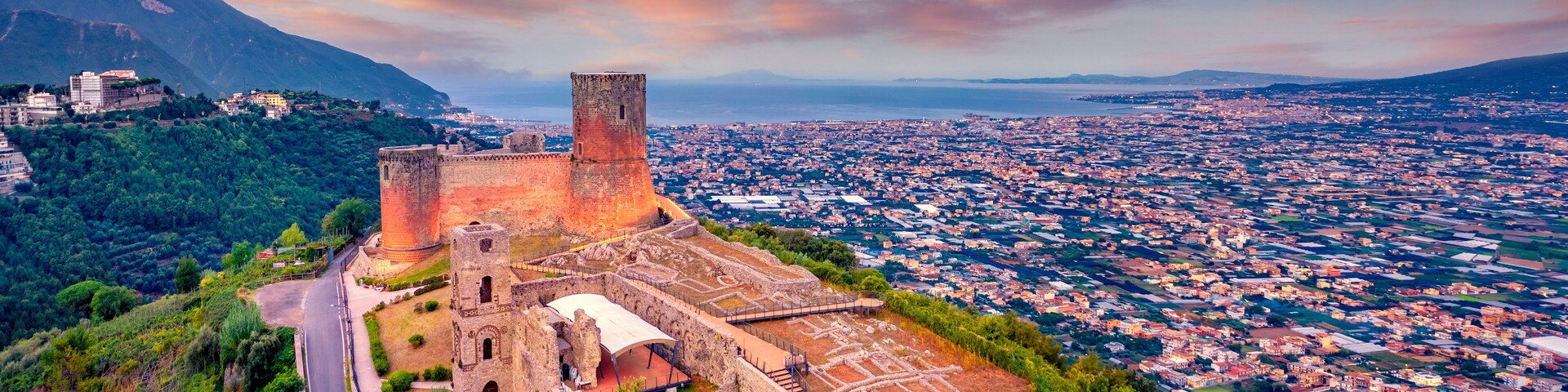 Uma fortaleza histórica com uma torre e ruínas, rodeada por vegetação exuberante e com vista para uma paisagem urbana costeira.