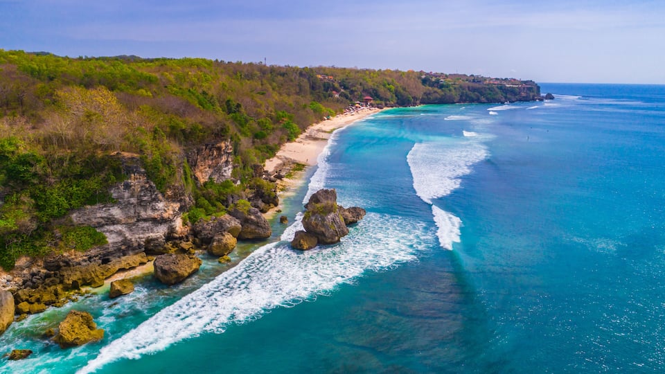 A coastal landscape with a clear blue sea, rocky outcrops, and a sandy beach.