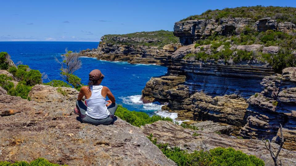 A person meditating on a rocky cliff overlooking a coastal area with cliffs and greenery.