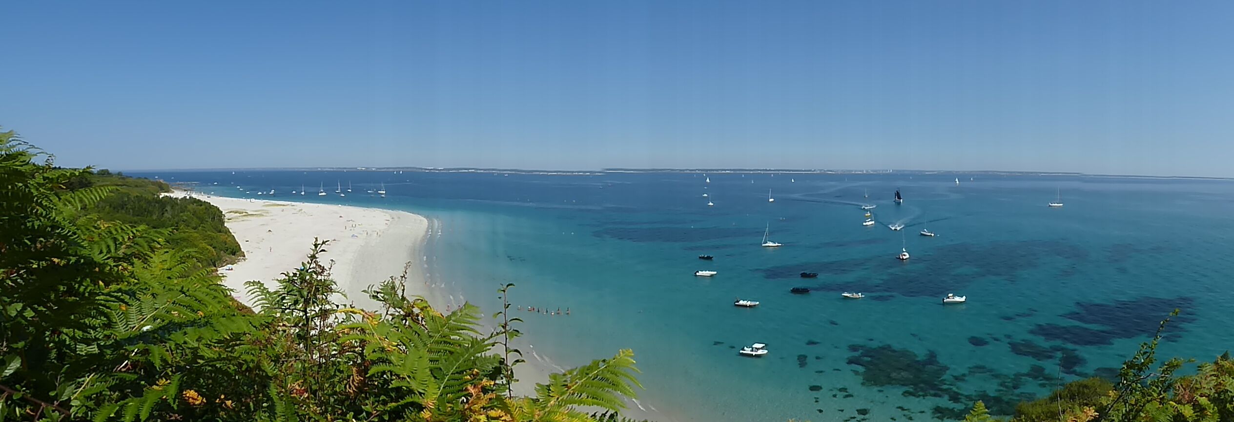 A coastal landscape with a sandy beach, clear turquoise waters, and numerous boats.