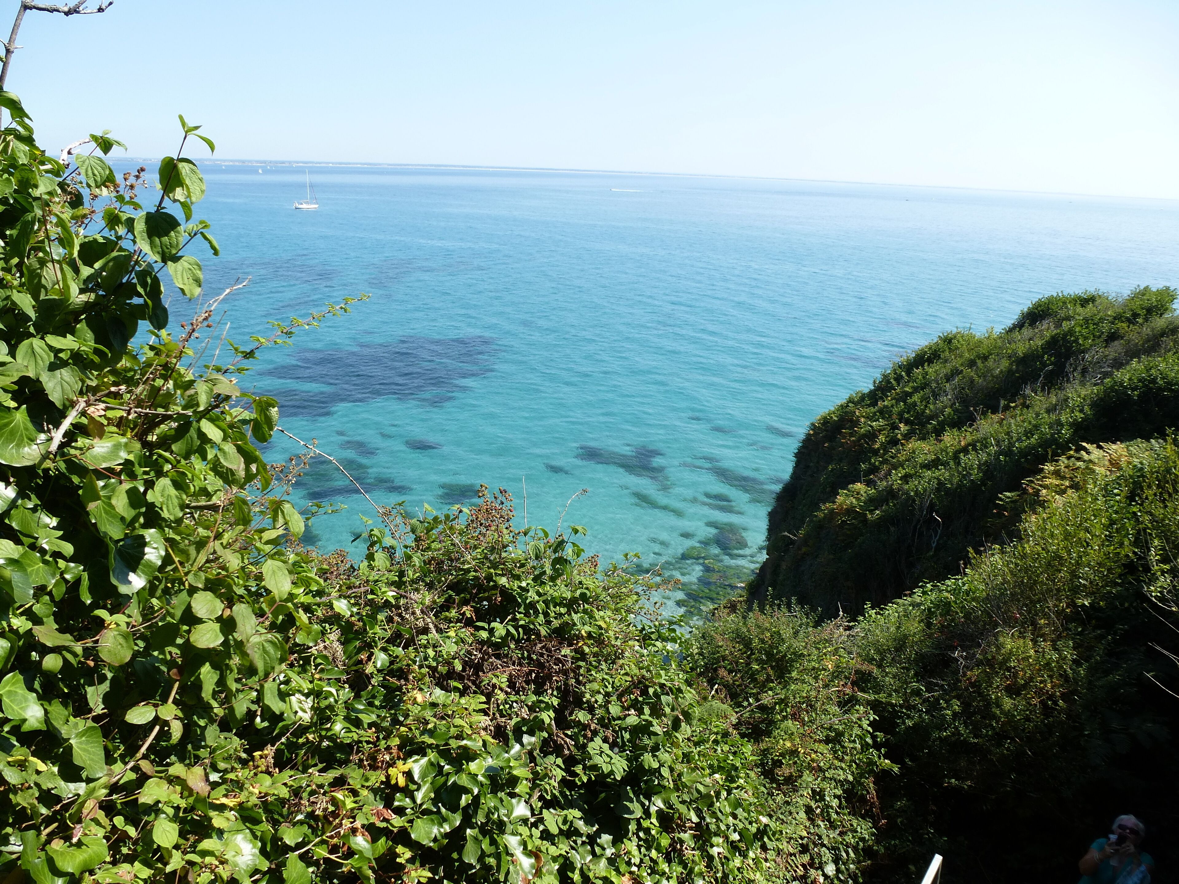 A coastal landscape with clear blue waters, a distant sailboat, and lush greenery.
