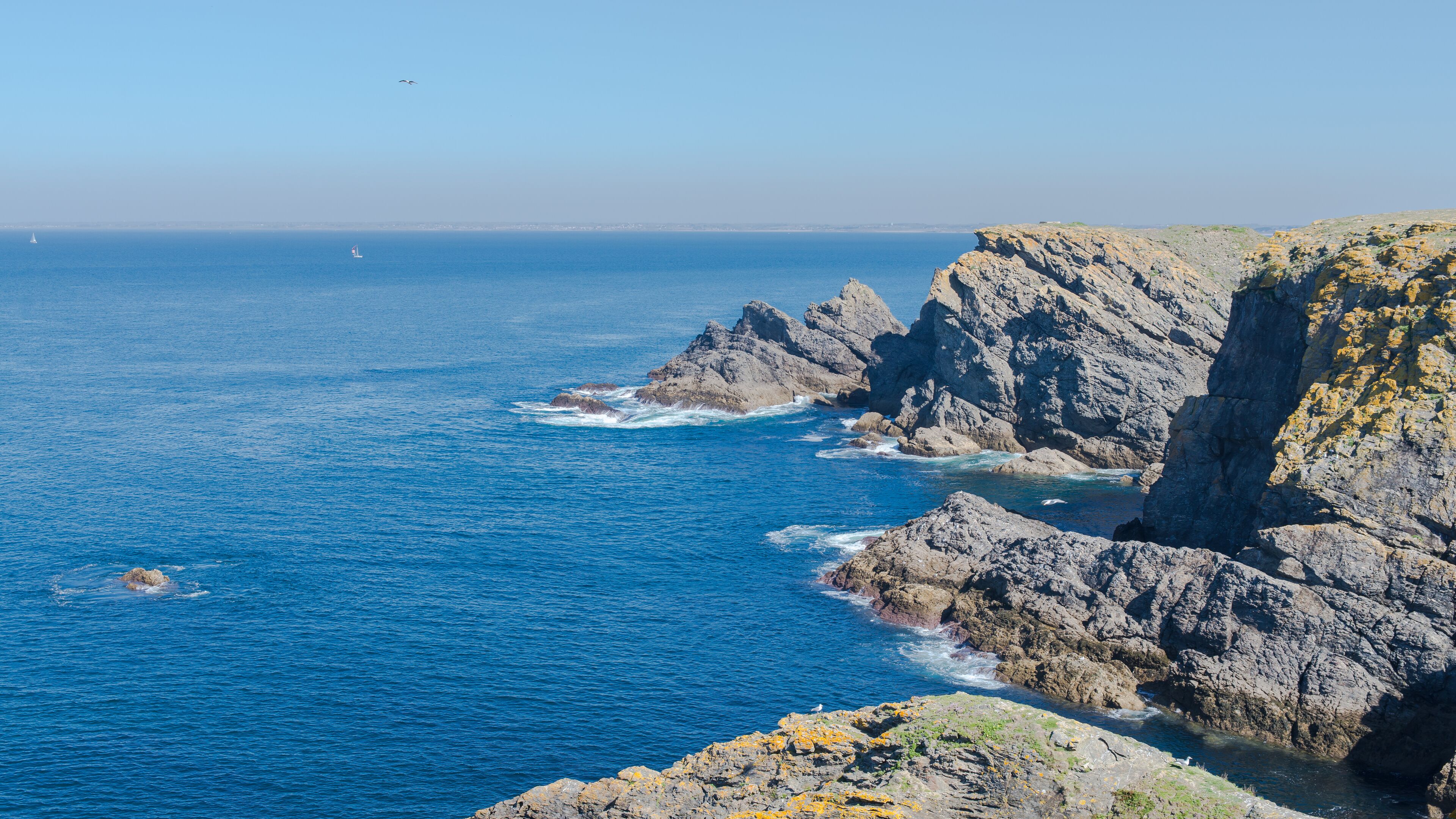 A rocky coastline with clear blue waters and a sailboat in the distance.