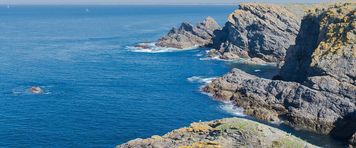 A rocky coastline with clear blue waters and a sailboat in the distance.