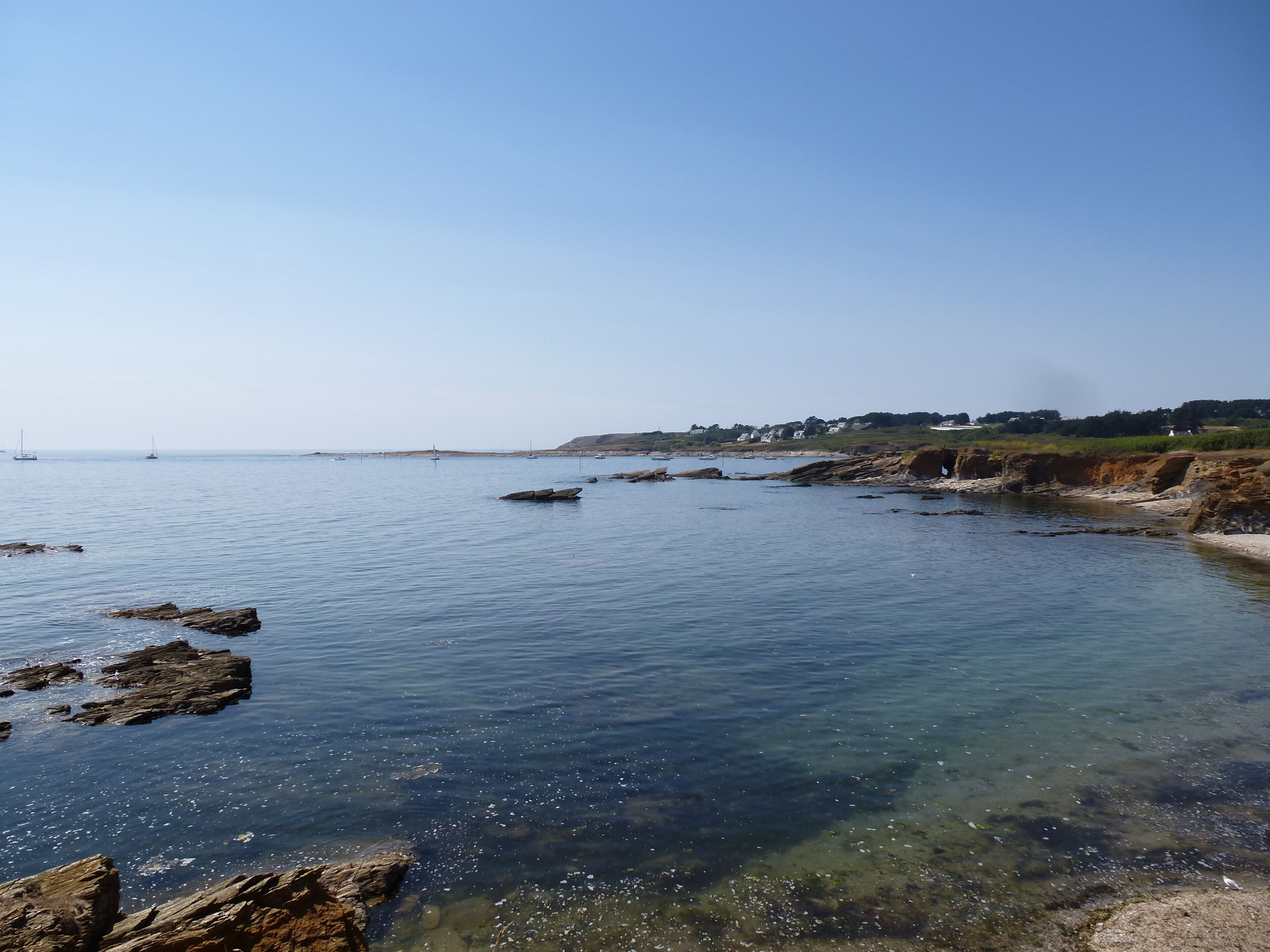 A coastal landscape with clear water, rocky shorelines, and sailboats in the distance.