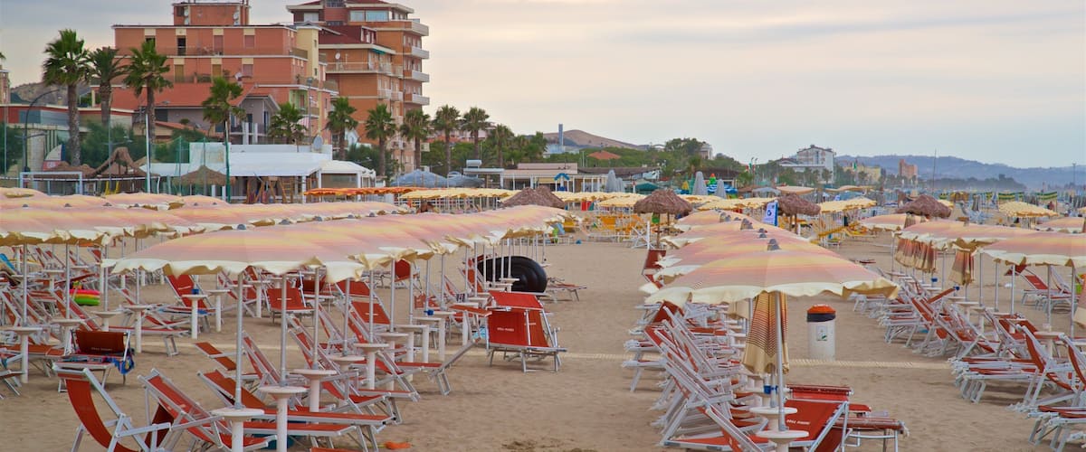 A beach with rows of sun loungers and umbrellas, a sandy shore, and buildings in the background.
