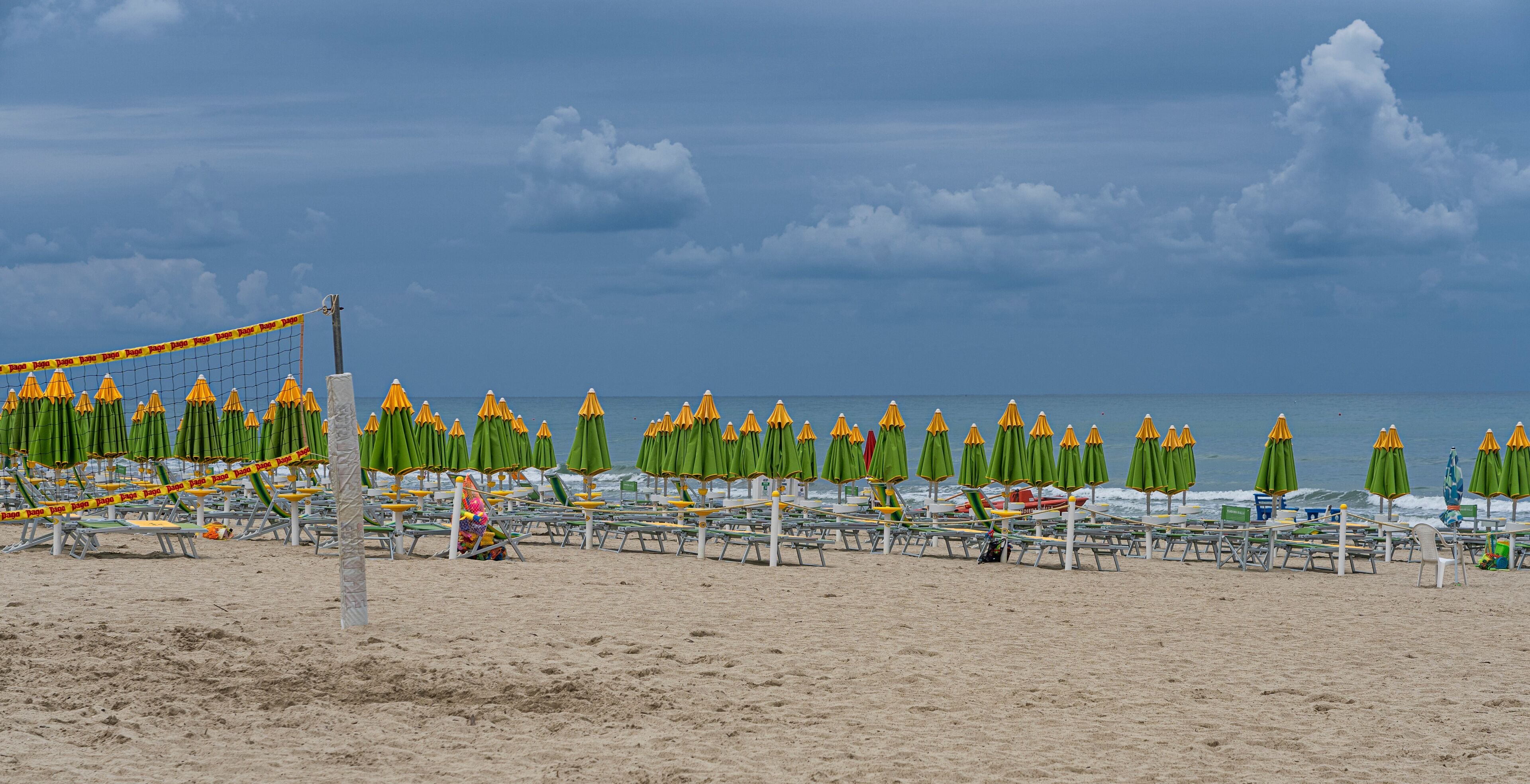 A beach with rows of green and yellow umbrellas, a volleyball net, and a sandy shore.