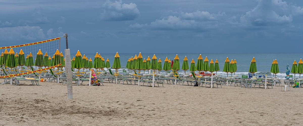 Una playa con hileras de sombrillas verdes y amarillas, una red de voleibol y una orilla arenosa.