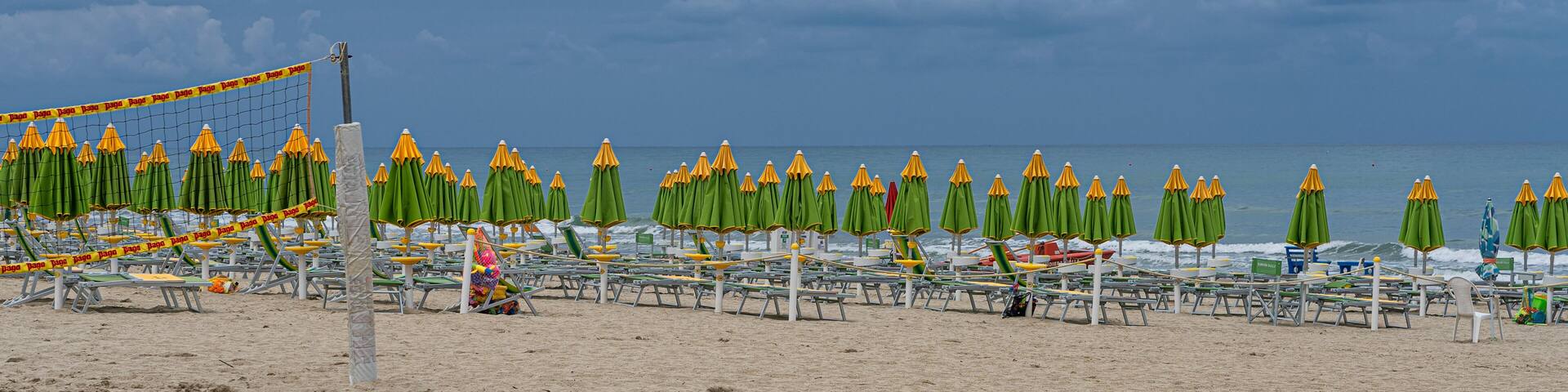 A beach with rows of green and yellow umbrellas, a volleyball net, and a sandy shore.