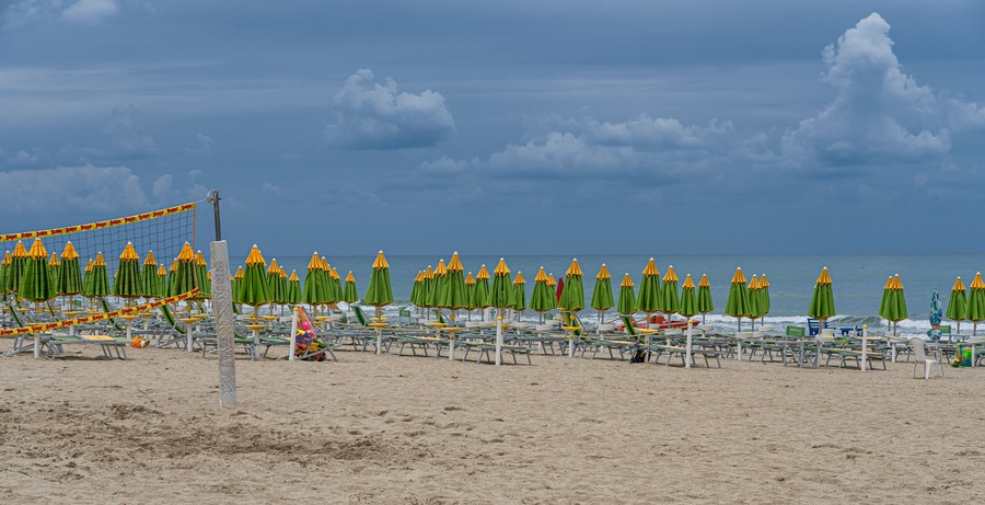 Une plage bordée de rangées de parasols verts et jaunes, avec un filet de volley-ball et une étendue de sable.