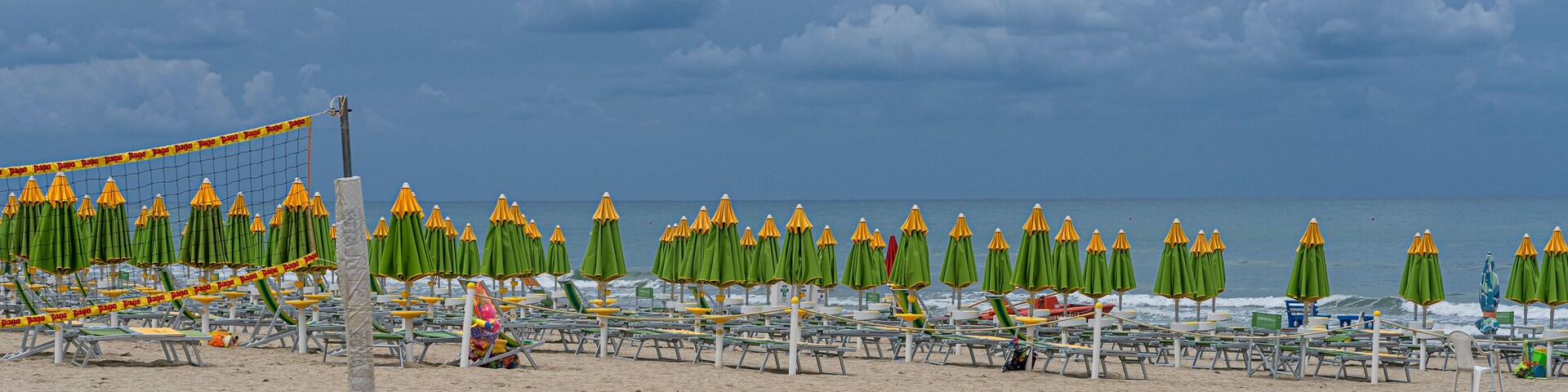Una playa con hileras de sombrillas verdes y amarillas, una red de voleibol y una orilla arenosa.