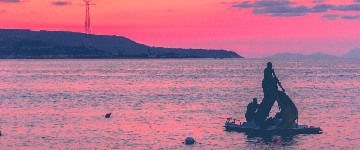 A sunset over a body of water with silhouettes of people on a boat and a distant power line tower.