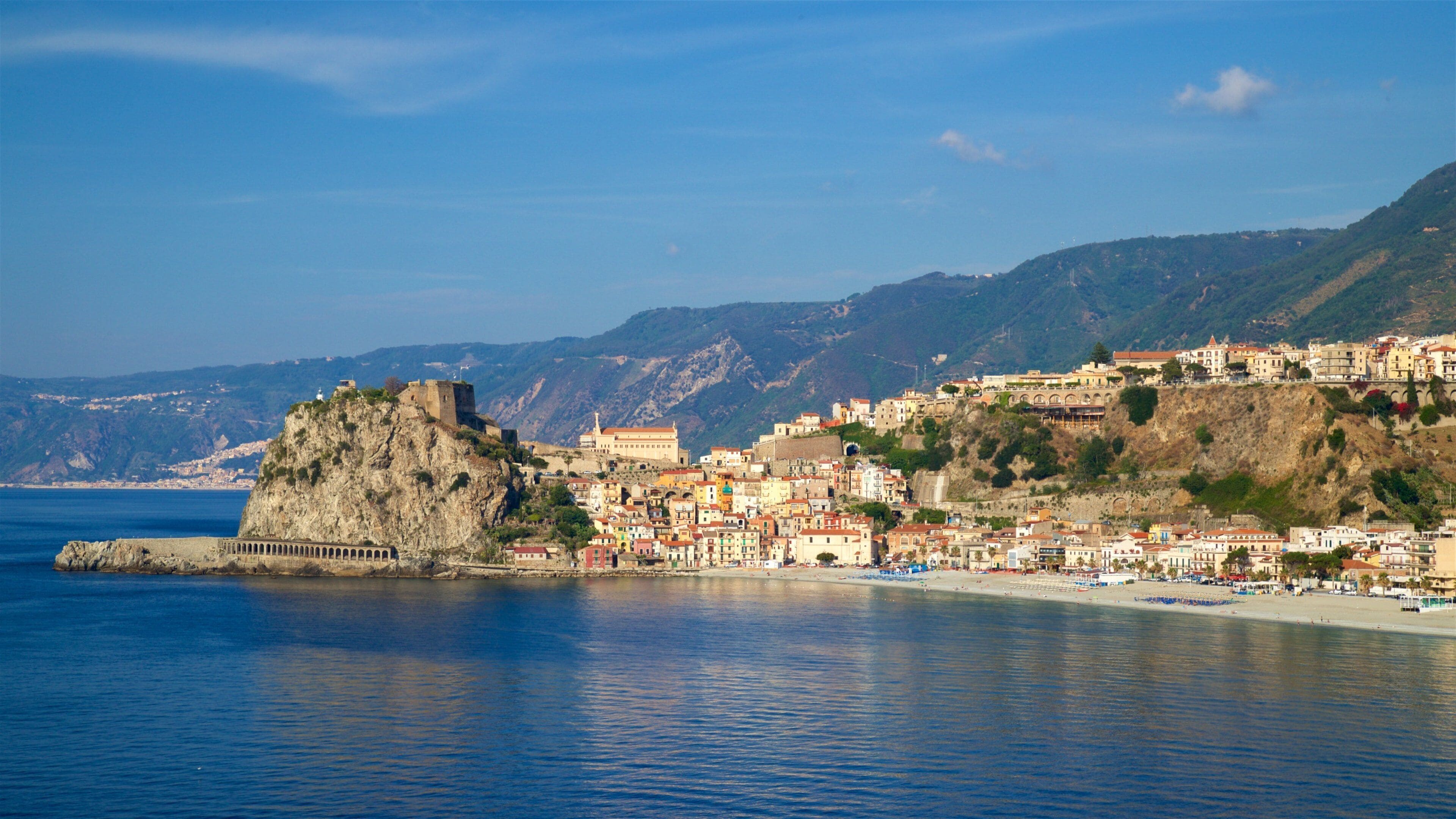 A coastal town with a prominent castle on a cliff, a beach, and a mountainous backdrop.