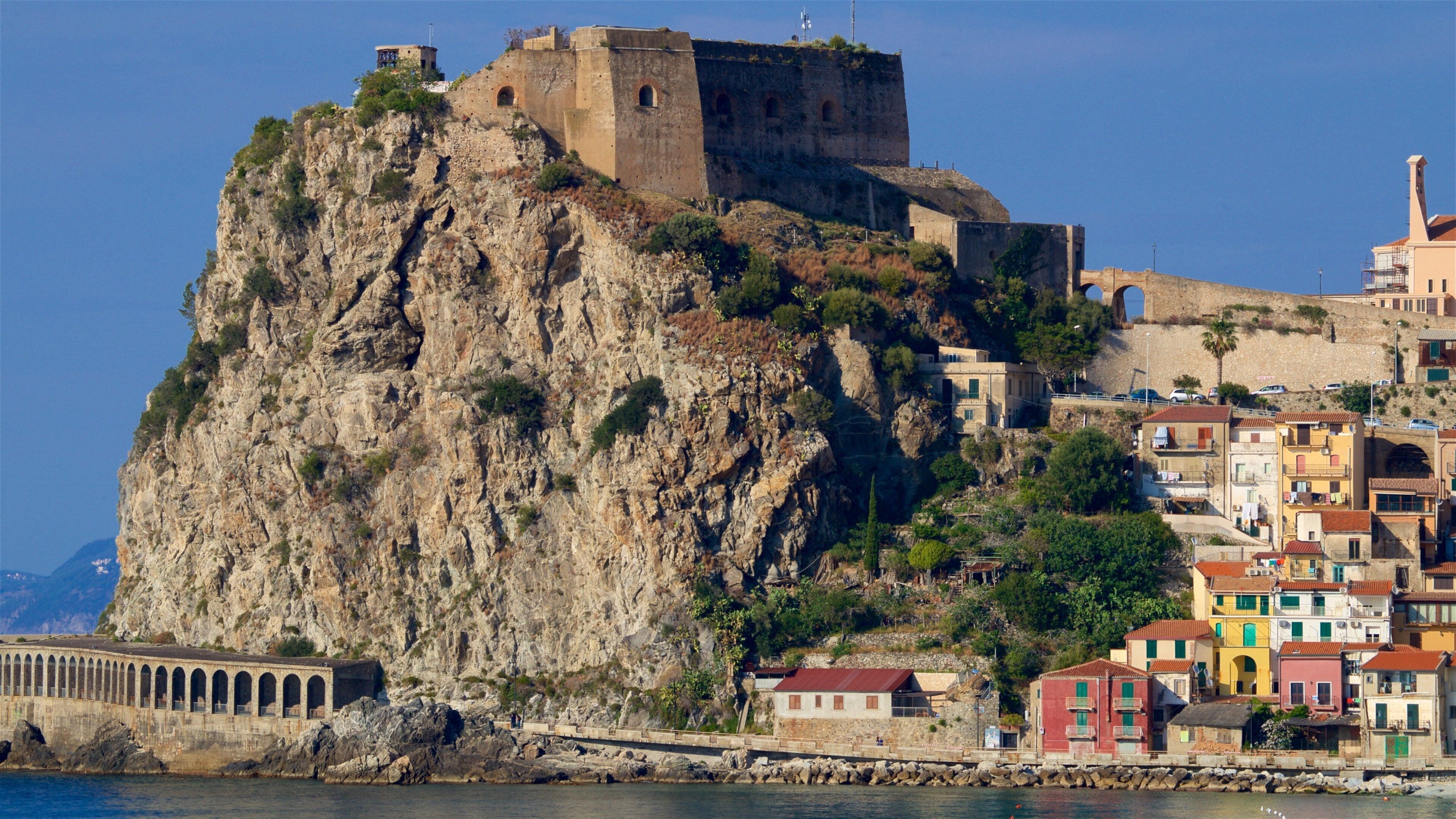 A coastal town with a prominent castle on a rocky cliff, colorful buildings, and a stone bridge.