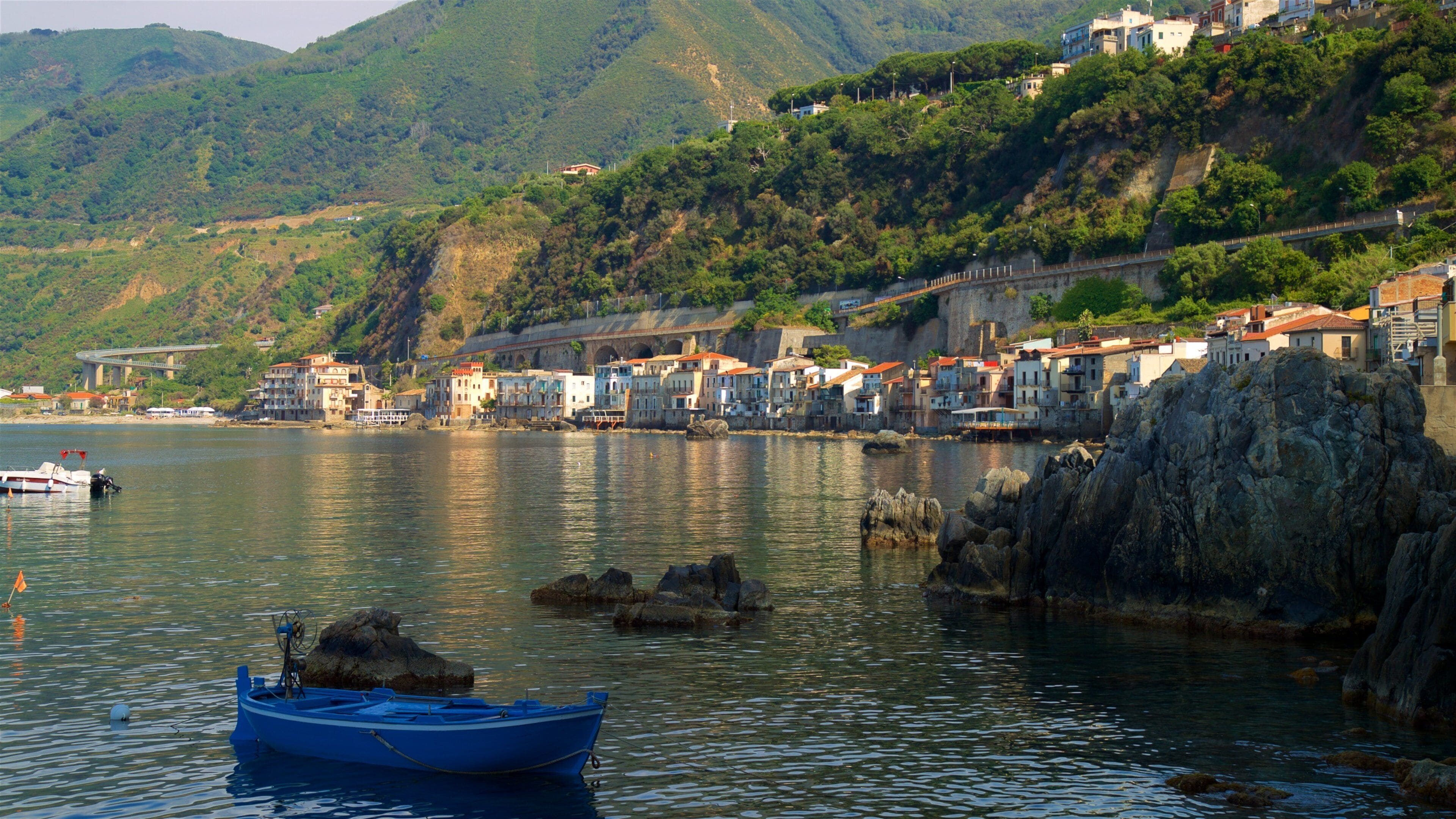 A coastal town with buildings on a hillside, a bridge, and a blue boat in the water.