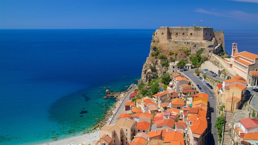 Un pueblo costero con una fortaleza histórica que domina el mar, edificios con tejados de teja y un océano azul cristalino.