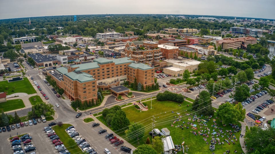 An aerial view of a university campus with multiple buildings, parking lots, and a large green field with people gathered.