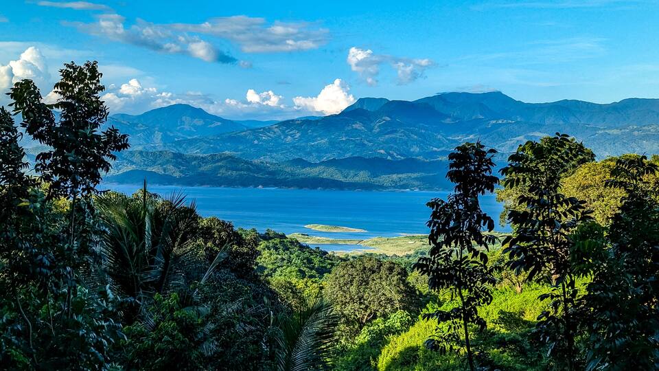 A coastal landscape with mountains in the background and dense greenery in the foreground.
