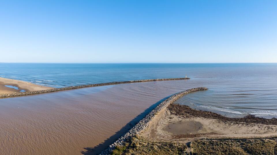 A coastal landscape with a curved breakwater, sandy beach, and calm sea.
