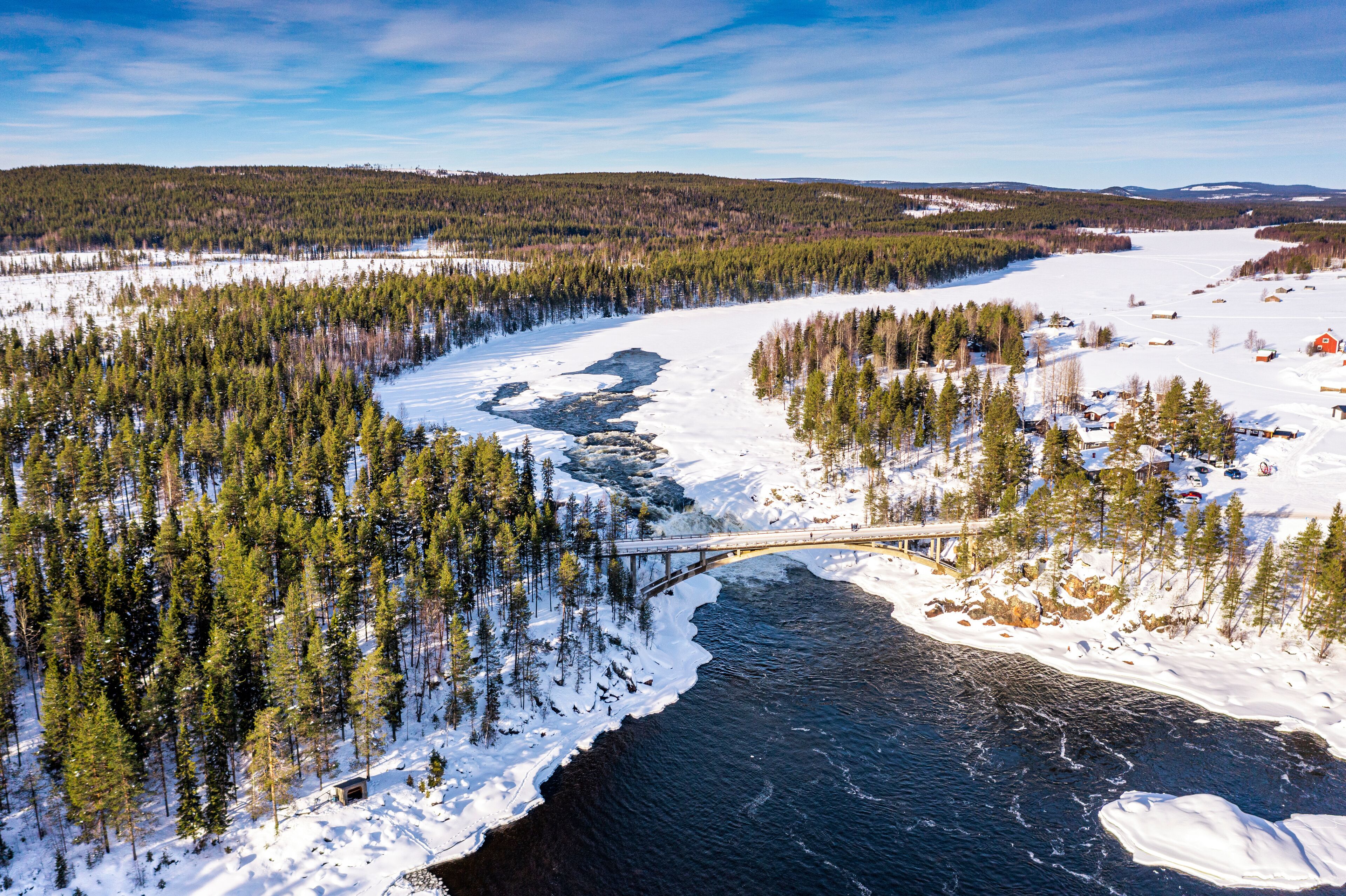 A snow-covered river with a bridge, surrounded by dense forests and a few buildings.