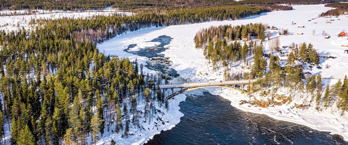 A snow-covered river with a bridge, surrounded by dense forests and a few buildings.