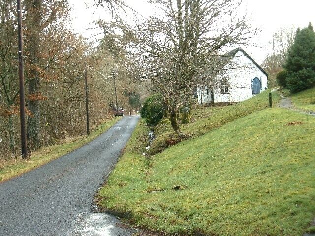 A rural road with a white building on the right, surrounded by trees and grass.