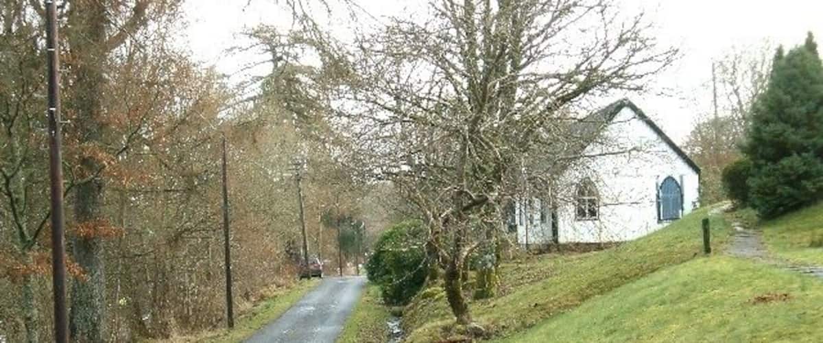 A rural road with a white building on the right, surrounded by trees and grass.